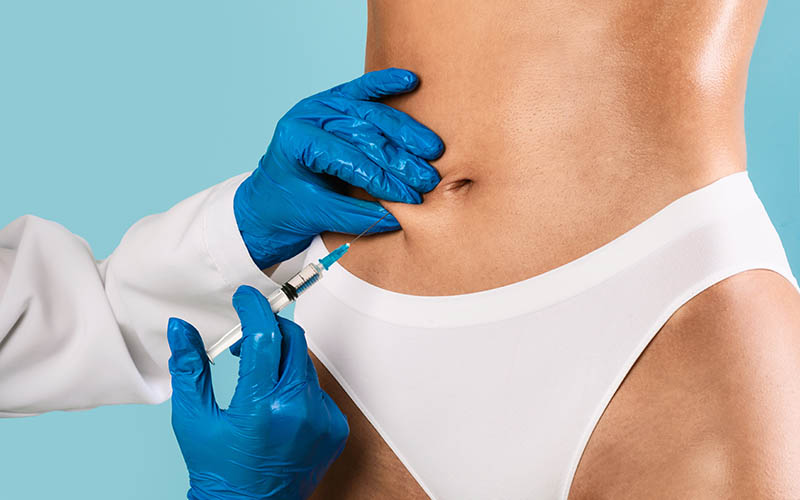 Plastic surgeon in protective gloves making injection at belly area for unrecognizable lady, blue studio background. Slim woman having lipolysis treatment at beauty salon, cropped close up view