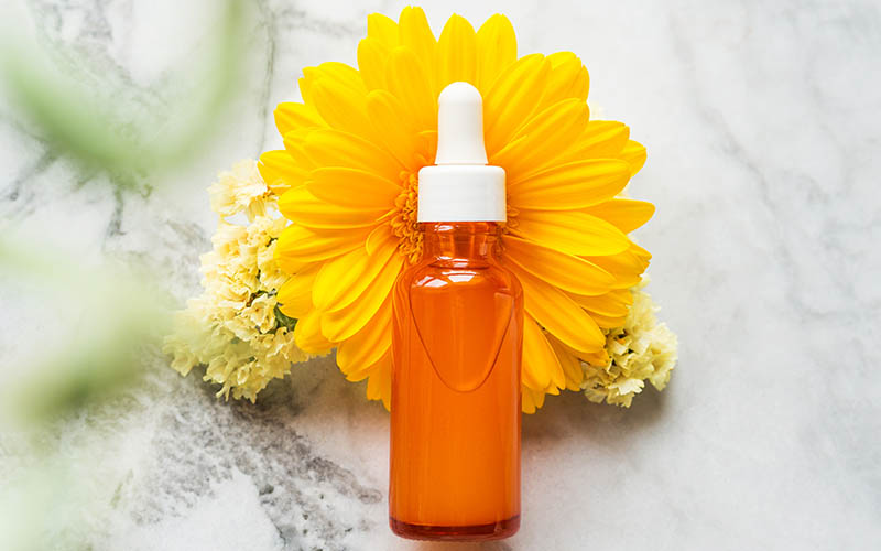 Skin care Vitamin C serum in orange bottle with dropper over beautiful yellow flowers on marble table. Natural anti aging beauty product. Top view