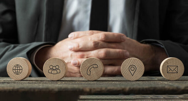 Wide view image of business customer service representative sitting at a desk with a line of wooden cut circles with contact and information icons on them in front of him.