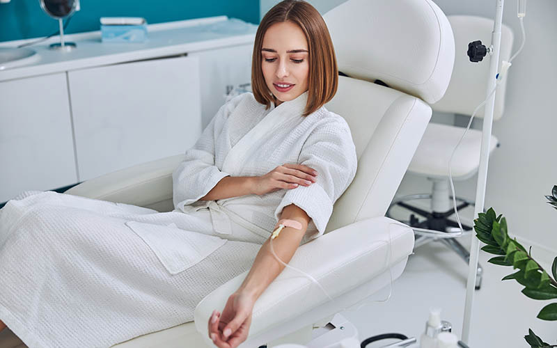 Waist up side view portrait of redheaded Caucasian female in white bathrobe sitting in medicine armchair during medical procedure in wellness center